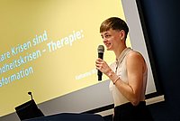 Katharina Wabnitz steht auf der Bühne vor einem gelben Hintergrund und hält ein Mikrofon in der Hand.